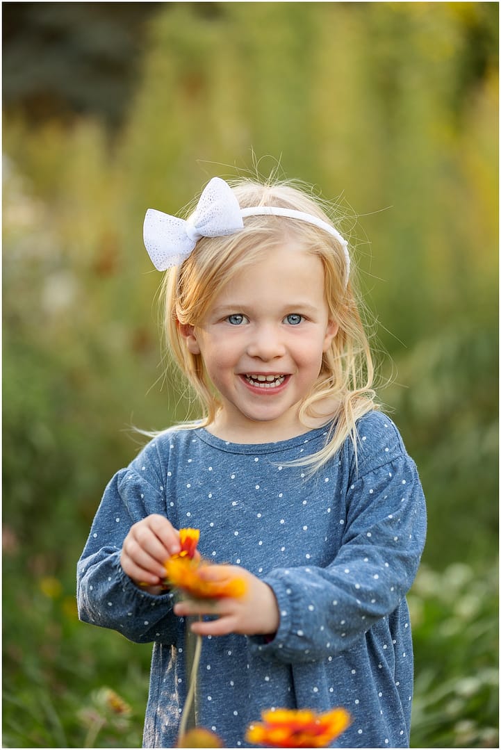 Little blonde girl in blue dress and white headband smiles big. Photo by Tiffany Hix Photography.