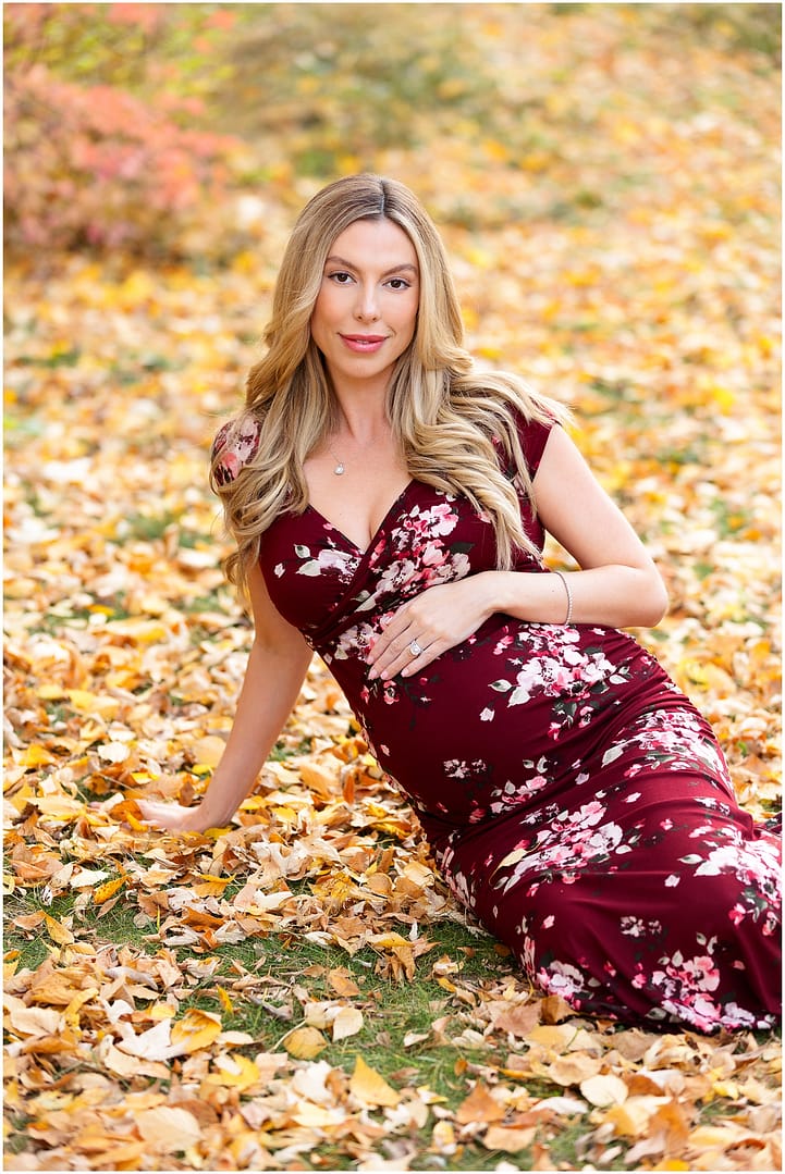 Mom sits down among fall leaves holding her baby bump. Photo by Tiffany Hix Photography.