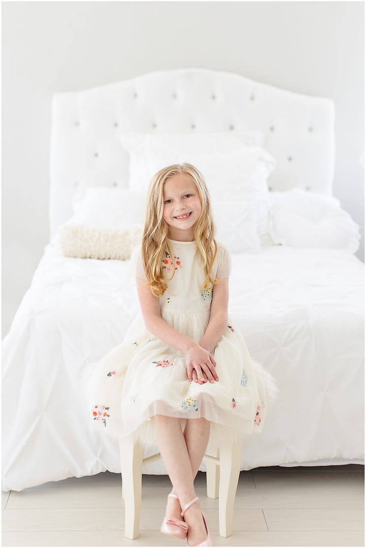 Little girl sits in spring dress in front of white bed. Photo by Tiffany Hix Photography.