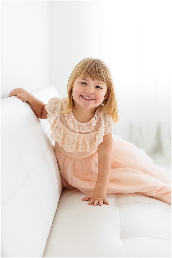 Little girl smiles while sitting on white couch during Boise childhood session. Photo by Tiffany Hix Photography.