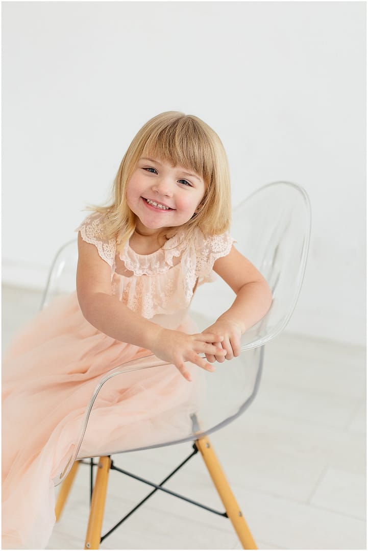 Little girl smiles while sitting in acrylic chair. Photo by Tiffany Hix Photography.
