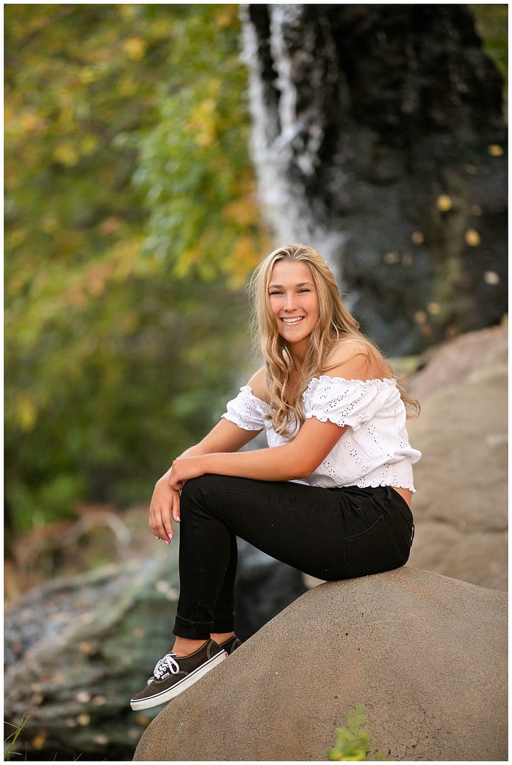 Blonde teen sits on rock in front of waterfall. Photo by Tiffany Hix Photography.