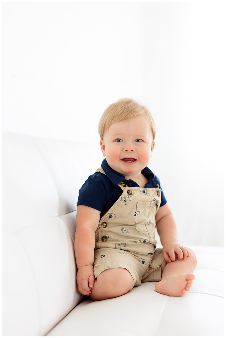 Baby boy in overalls sits and smiles during milestone session. Photo by Tiffany Hix Photography.